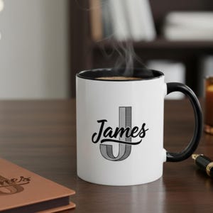 Puede incluir: Taza de cerámica blanca con asa y borde negros. La taza presenta el nombre "James" en una escritura elegante con una gran inicial "J" a rayas. Sale vapor de la taza, lo que sugiere una bebida caliente.