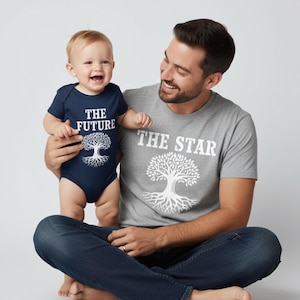 Puede incluir: Un bebé con un body azul marino con el texto "THE FUTURE" y un gráfico de árbol es sostenido por una persona que lleva una camiseta gris con "THE STAR" y un gráfico de árbol a juego. Ambos sonríen.