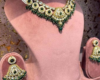Puede incluir: Un conjunto de collar y pendientes en tono dorado con hilos de perlas blancas, detalles de piedras verdes circulares y piedras transparentes incrustadas. La joyería presenta pequeños colgantes de cuentas verdes en los bordes inferiores del collar y los pendientes triangulares.