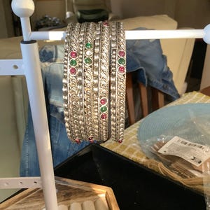 May include: A set of four silver-toned metal bangles featuring a twisted rope-like texture and inlaid with alternating round red and green stones. The bangles are displayed hanging on a white metal jewelry stand.