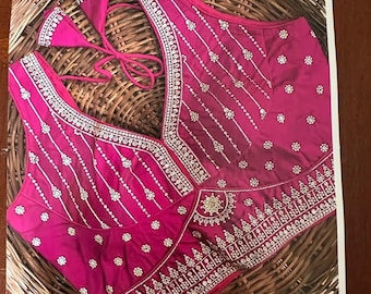 Puede incluir: Una blusa fucsia sin mangas con un escote en V profundo, intrincados bordados en blanco y plata, y pequeños motivos florales. La prenda incluye un cierre de lazo en la espalda con dos borlas decorativas y un borde inferior bordado.