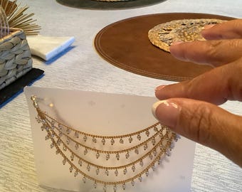 Puede incluir: Un collar de varias cadenas doradas en cascada. Cada cadena está adornada con pequeñas cuentas blancas espaciadas uniformemente que cuelgan de los eslabones. El collar se presenta sobre una tarjeta blanca rectangular para su exhibición.