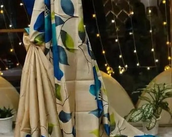 Può includere: Un sari beige caratterizzato da grandi fiori blu dipinti a mano con foglie verdi e sottili steli neri. Il tessuto ha una lucentezza sottile e il motivo floreale è distribuito lungo tutta la lunghezza del capo, mettendo in risalto petali blu.