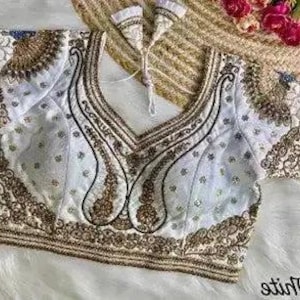 May include: A white and gold embroidered blouse with a V-neck design. The blouse features intricate gold embroidery along the neckline, sleeves, and hem. The sleeves are short and the fabric appears to be satin or a similar material. The blouse is likely part of a traditional or formal outfit.
