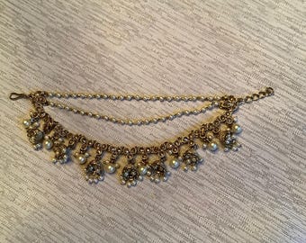 Puede incluir: Un adorno para la cabeza de metal dorado con un diseño de cadena de doble capa adornado con pequeñas perlas blancas. La sección inferior consta de una banda decorativa con dijes colgantes en forma de campana, acentuados con perlas blancas.