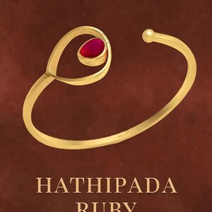May include: A gold-toned open cuff bracelet featuring a single teardrop-shaped red ruby gemstone set within a swirling loop design. The bracelet has a small spherical bead at the open end. The text HATHIPADA RUBY is printed below the jewelry piece.