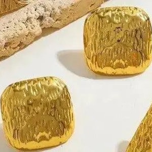 May include: A set of square-shaped gold-colored buttons with a textured, hammered surface. Each button features a rounded square silhouette with a reflective, metallic finish and irregular indentations across the top surface.