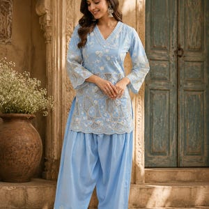 May include: A light blue tunic and matching loose-fitting trousers set. The tunic features a V-neckline, three-quarter length sleeves, and intricate white floral embroidery with geometric cut-out patterns along the hem and mid-section.
