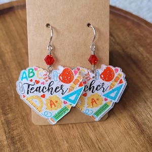 May include: A pair of teacher-themed earrings. Each earring is heart-shaped with the word "Teacher" and school-related graphics. The earrings have silver hooks and red bead accents.