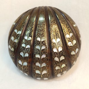 Op de afbeelding: Een ronde houten doos met een donkerbruine afwerking. De doos is versierd met gouden filigraan en parelmoer inlegwerk. Het ontwerp heeft verticale panelen met een herhalend bladpatroon.