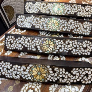 May include: Four rectangular dark wood blocks featuring intricate mother-of-pearl inlay. Each block displays a central floral medallion surrounded by swirling white shell patterns, set against a dark, polished wooden surface with gold accents.