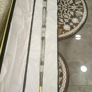 Pode incluir: Uma bengala ornamentada com uma pega preta e um eixo decorativo branco e dourado. A bengala é apresentada numa caixa forrada a cetim branco com acabamentos a preto e dourado. O eixo da bengala apresenta padrões e desenhos intrincados.