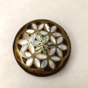 May include: A round, decorative object with a floral design. The object features white, iridescent, and dark brown elements. The outer edge is a dark brown color, and the center has a mother-of-pearl appearance.