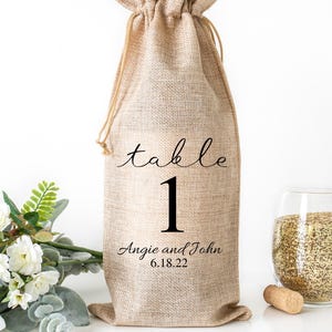 May include: A beige burlap wine bottle bag with a drawstring closure. The bag is printed with the word "table", the number "1", the names "Angie and John", and the date "6.18.22" in black script. A glass of seeds and a cork are next to the bag.