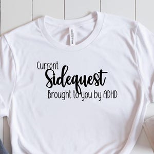 May include: White t-shirt with the text "Current Sidequest Brought to you by ADHD" in black script. The shirt is paired with gray jeans and black canvas sneakers with white laces. A book and a cup of coffee are also visible.