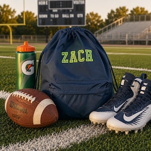 May include: A football-themed collection featuring a brown Wilson football, a green and orange water bottle, a navy blue drawstring backpack with "ZACH" in green, and navy blue and white football cleats on a green field.