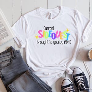 May include: White t-shirt with the text "Current Sidequest Brought to you by ADHD" in colorful letters. The shirt is paired with gray jeans and black and white sneakers. A book and a cup of coffee are also in the image.