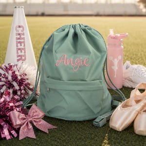 May include: A sage green drawstring backpack featuring the name Angie embroidered in pink script. The bag includes a front zippered pocket and matching green cord straps. It is surrounded by a white megaphone, pink water bottle, and pom-poms.