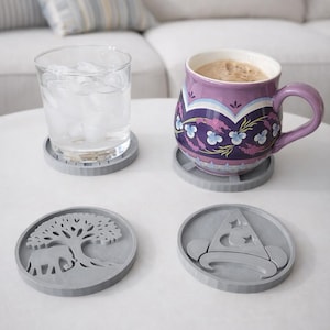 May include: A purple floral mug with coffee sits on a gray coaster, next to a glass of water. Two additional gray coasters feature an elephant and a wizard hat design. The coasters are on a white table.