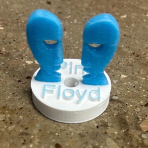 Może przedstawiać: Niebieska rzeźba 3D z dwoma stylizowanymi twarzami zwróconymi do siebie, zamontowana na białej, okrągłej podstawie. Podstawa ma napis "Pink Floyd" w wypukłych literach. Rzeźba znajduje się na nakrapianej powierzchni.