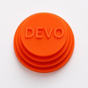 Puede incluir: Objeto circular, en capas, de color naranja brillante con la palabra "DEVO" en letras en relieve. El objeto tiene una serie de círculos concéntricos, cada uno ligeramente más pequeño que el anterior, creando un efecto escalonado. El fondo es blanco liso.