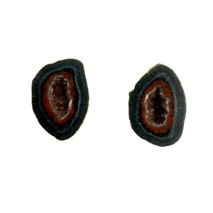 May include: A pair of oval earrings with a geode design. Each earring has a dark grey outer edge, a red-brown inner layer, and a light brown centre, resembling a miniature geode.