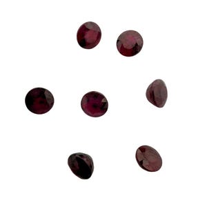 May include: Seven oval-cut, deep red gemstones are scattered against a white background. The gems have a faceted surface, catching the light and displaying a rich, dark color. These gemstones could be used for jewelry making.