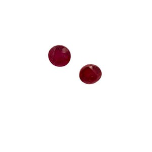 Puede incluir: Dos gemas redondas, facetadas, de color rojo intenso, posiblemente rubíes, se muestran sobre un fondo blanco. Las gemas tienen una superficie pulida y reflectante, y están ligeramente separadas.