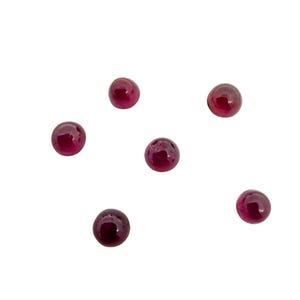 May include: Six small, round, polished gemstones in a deep red color are scattered against a white background. The gems have a glossy finish, reflecting light. These could be used for jewelry making or other crafts.