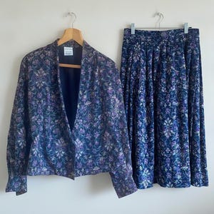 May include: A two-piece set consisting of a long-sleeved jacket and a pleated skirt, both featuring a dark blue fabric with an intricate floral pattern in shades of purple, light blue, and white. The jacket has a simple open front design.