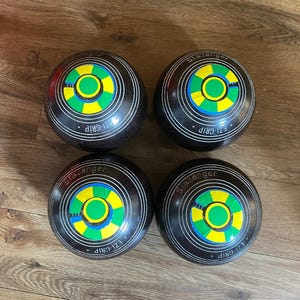 May include: Four black lawn bowls featuring white concentric rings and a central circular emblem with green and yellow segments. Each bowl is marked with the text Slazenger and EZI-GRIP in white lettering on the dark, spherical surface.