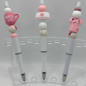 Pode incluir: Três canetas brancas com topos decorativos. Uma caneta tem uma caneca rosa que diz "NURSE LIFE". Outra tem um chapéu de enfermeira com uma cruz rosa. A terceira caneta tem um uniforme de enfermeira rosa. Todas as canetas têm detalhes prateados.