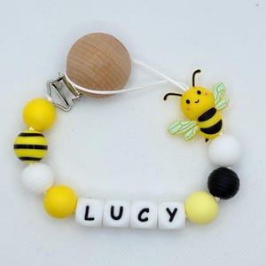 Op de afbeelding: Een fopspeenclip voor baby's met een houten clip en een houten kraal. Het heeft gele, zwarte en witte kralen en een gele bijenbedel. De naam "LUCY" is gespeld met witte letterkralen.