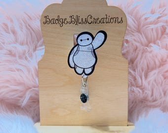 May include: A badge reel featuring a cartoon character with a white body, black outline, and waving arm. The reel is attached to a wooden display stand with the text "Badge Bliss Creations". The background is a soft, pink, fluffy material.
