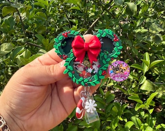 Puede incluir: Un portatarjetas circular con diseño de corona verde y lazo rojo, relleno de confeti navideño. Un dije de bastón de caramelo y un dije de copo de nieve blanco cuelgan de la parte inferior. Una pegatina con un logotipo está en el lateral.