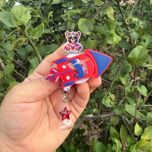 May include: A red, white, and blue rocket-shaped badge reel with a star-shaped accent. The reel features a clear plastic body with a rocket design and a retractable cord. The badge reel has the text "Badge Bliss Creations".