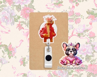 Puede incluir: Un portatarjetas con un personaje de dibujos animados con una chaqueta roja y un perro de dibujos animados con un vestido rosa y una corona. El carrete está unido a una tarjeta rectangular marrón. El fondo es un estampado floral.