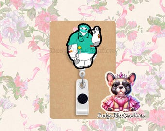 May include: A retractable badge reel featuring a cartoon character in medical scrubs, a smiling face, and a small cartoon dog wearing a pink dress and crown. The text "Badge Bliss Creations" is visible.