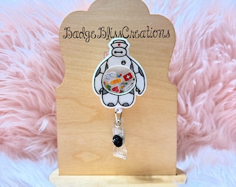 May include: A wooden badge holder with the text "Badge Bliss Creations" features a white and black cartoon character with a nurse's hat. The character's body contains various pills and medical supplies. A clear badge reel with a black button is attached.