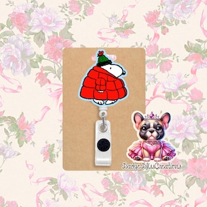 May include: A Snoopy badge reel with a red coat and green hat. A French Bulldog in a pink tutu and crown is also featured. The text "Badge Bliss Creations" is visible.