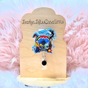 May include: A wooden badge holder with the text "Badge Bliss Creations" and a colorful badge featuring characters from a movie. The badge has a black button and a retractable reel. The background is a light pink, fluffy material.