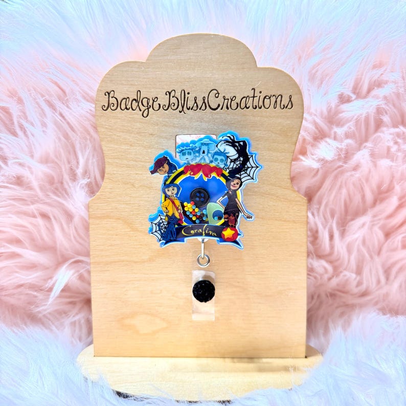 May include: A wooden badge holder with the text "Badge Bliss Creations" and a colorful badge featuring characters from a movie. The badge has a black button and a retractable reel. The background is a light pink, fluffy material.
