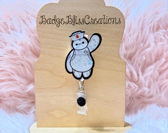 May include: A white and black badge reel featuring a cartoon character in a nurse's uniform. The character is waving with a red cross on the hat. The badge reel is attached to a clear plastic holder with a black button.