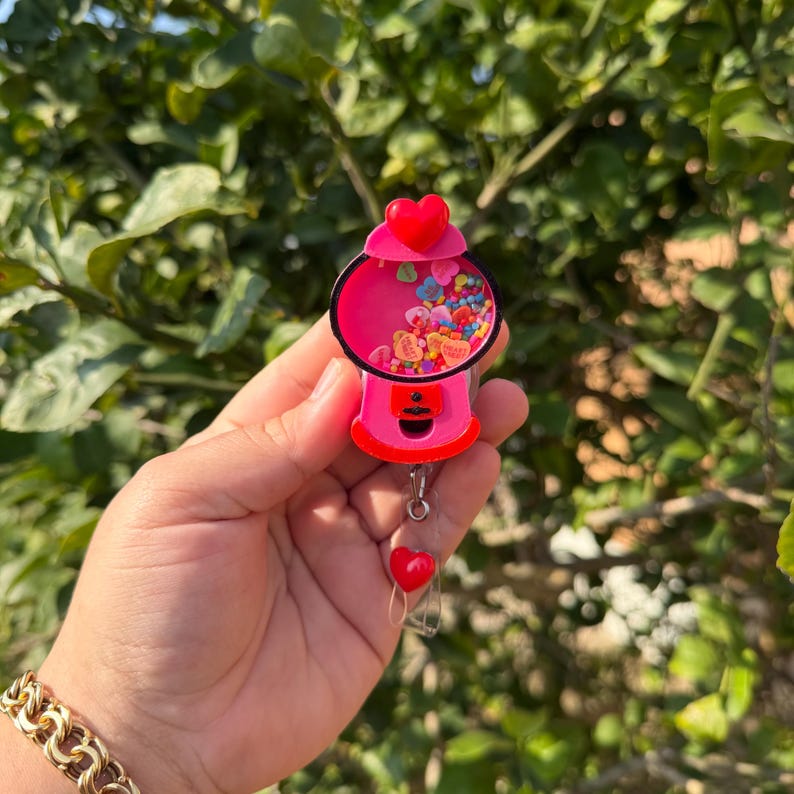May include: A pink gumball machine badge reel with a red heart on top and a dangling red heart charm. The gumball machine has a clear front filled with colorful heart-shaped confetti. The reel is held in a hand.