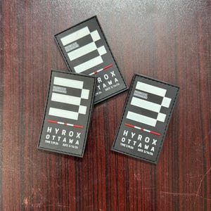 May include: Three rectangular black patches featuring a white geometric logo, a thin red and white striped line, and white text reading HYROX OTTAWA, TIME 1:19:34, and DATE 5/14/26. Each patch includes a small box with additional text details.