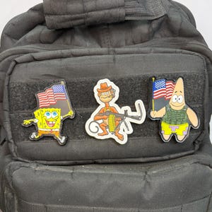 May include: A black tactical backpack featuring three morale patches attached to the front webbing. The patches depict a yellow sponge character holding an American flag, a brown monkey wearing a hat with a machine gun, and a pink starfish wearing a tactical vest and holding an American flag.