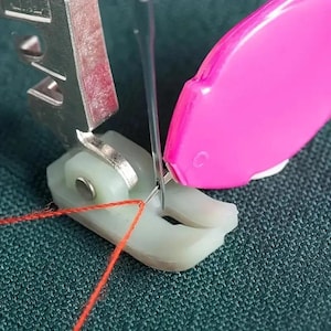May include: Close-up of a sewing machine needle piercing orange thread. The machine has a silver metal presser foot and a pink thread cutter. The fabric is a textured teal color.