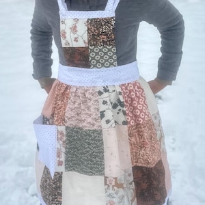 May include: A patchwork apron with a white polka dot waistband and pocket. The apron features squares of fabric in various patterns and colors, including floral, geometric, and solid designs. The straps are white with small polka dots.