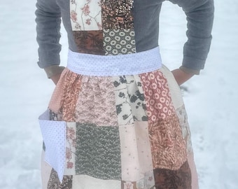 Könnte beinhalten: Eine Patchwork-Schürze mit weißem Pünktchen-Bund und Tasche. Die Schürze hat Stoffquadrate in verschiedenen Mustern und Farben, darunter Blumen-, geometrische und unifarbene Designs. Die Träger sind weiß mit kleinen Punkten.