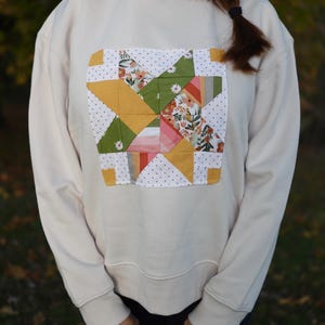 May include: Cream-colored sweatshirt featuring a patchwork design. The square patch showcases a starburst pattern with floral, geometric, and dotted fabric elements in shades of yellow, green, pink, and white. The sweatshirt has long sleeves and a crew neck.